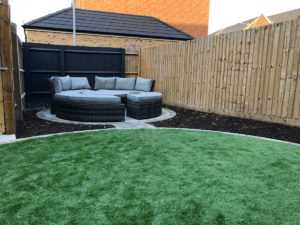 circular paved seating area in narrow garden with artificial grass in foreground