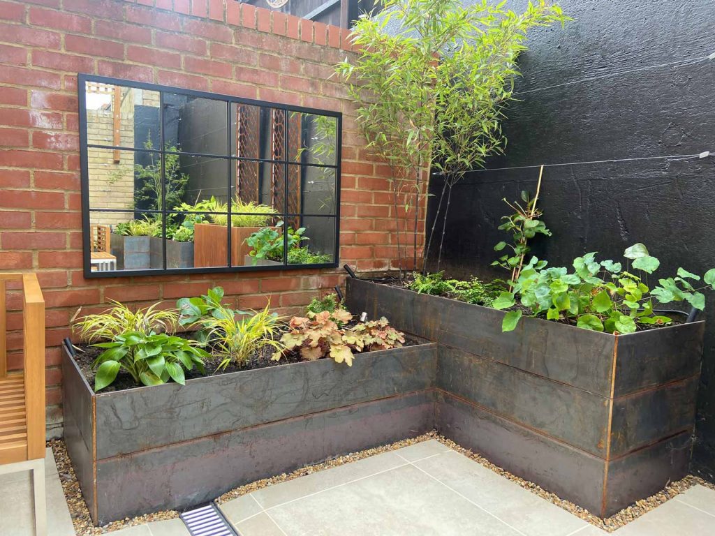 Two dark steel planters at right angles to each other with the rest of the garden reflected in an outdoor mirror